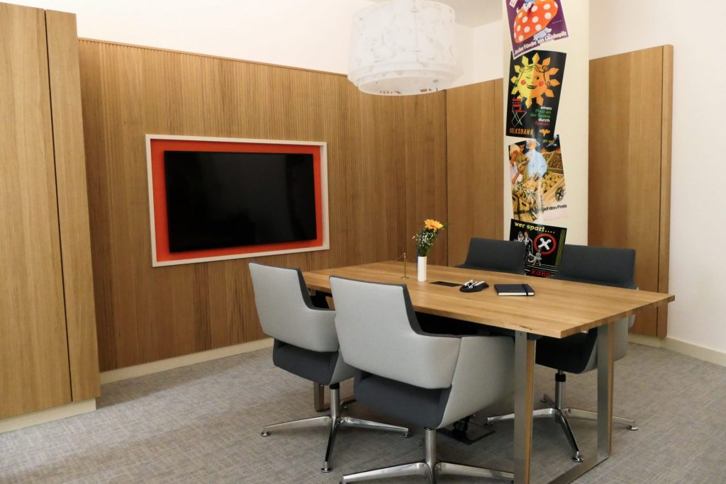 Konferenzraum mit elegantem Holztisch, vier Stühle und großem Wandmonitor, kombiniert moderne Technik mit warmem Holzambiente.