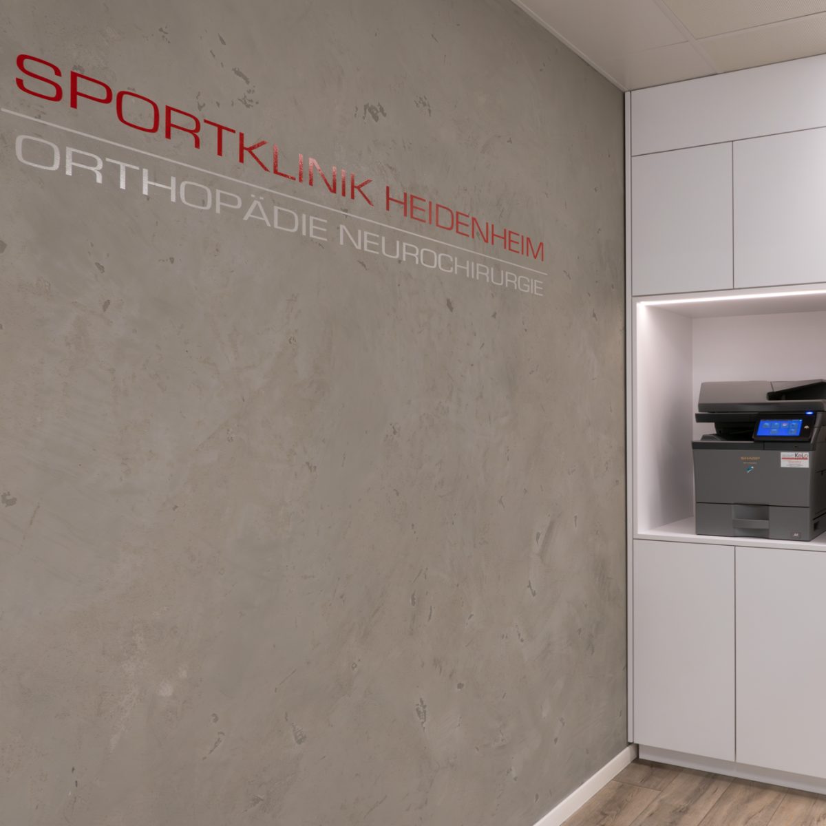 Innenaufnahme der Arztpraxis: Wand mit Praxisnamen und deckenhohem, beleuchtetem Einbauschrank, der einen Drucker enthält.