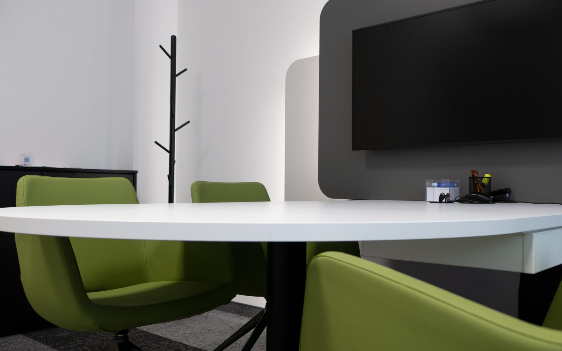 Helles, stilvolles Besprechungszimmer mit farbigen grünen Stühlen und weißem Tisch; die graue Wand mit montiertem Fernseher setzt einen modernen, hochwertigen Akzent.