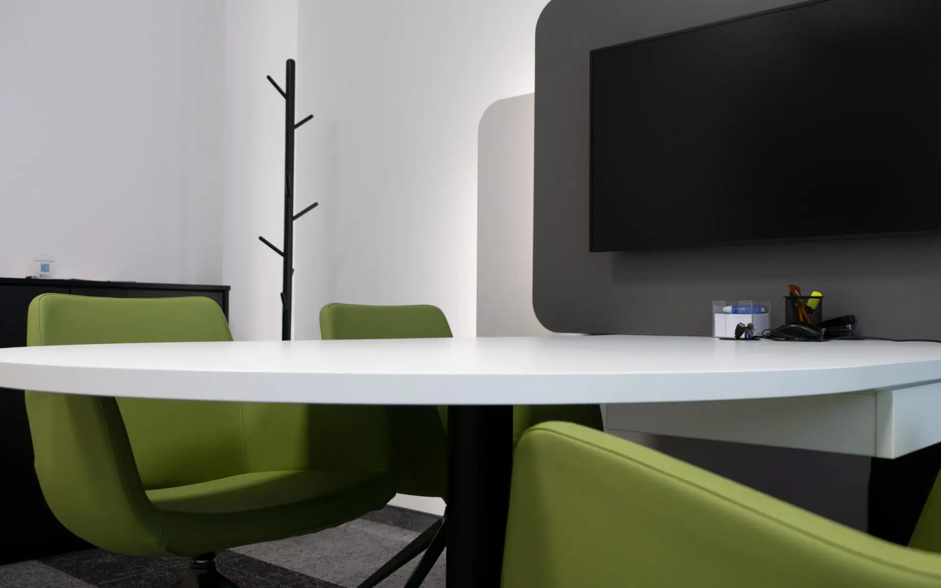 Helles, stilvolles Besprechungszimmer mit farbigen grünen Stühlen und weißem Tisch; die graue Wand mit montiertem Fernseher setzt einen modernen, hochwertigen Akzent.