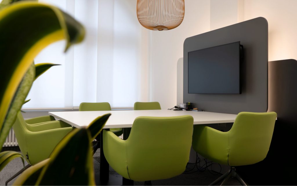 Modernes Besprechungszimmer einer Bank mit weißen Tischflächen, grünen Stühlen und einer grauen Akzentwand, an der ein Fernseher elegant integriert ist.