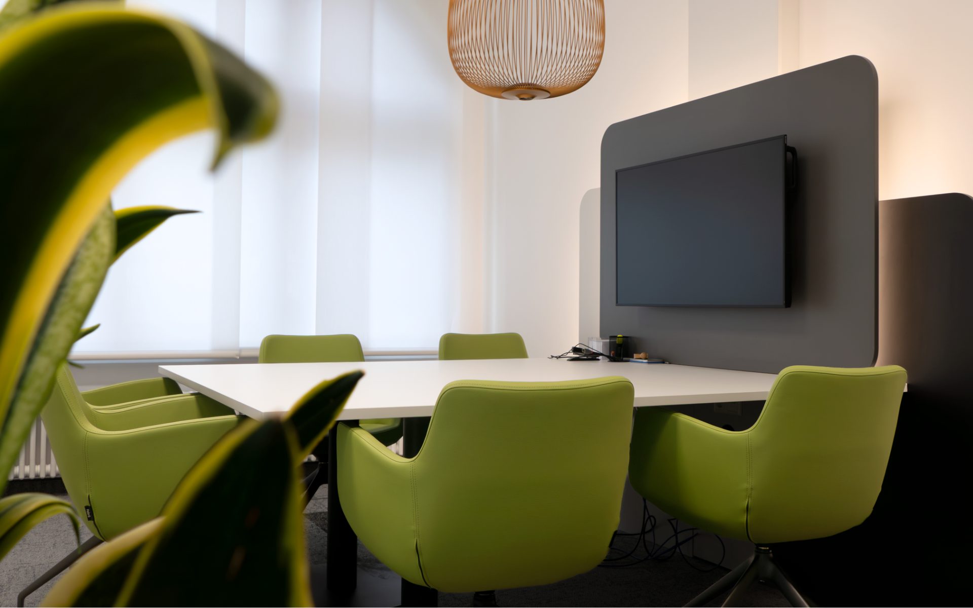 Modernes Besprechungszimmer einer Bank mit weißen Tischflächen, grünen Stühlen und einer grauen Akzentwand, an der ein Fernseher elegant integriert ist.