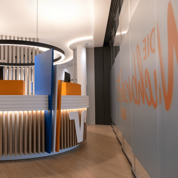 Moderner Empfangsbereich einer Raiffeisenbank mit runder Holzlamellen‑Theke und farblich abgestimmtem Sichtschutz.